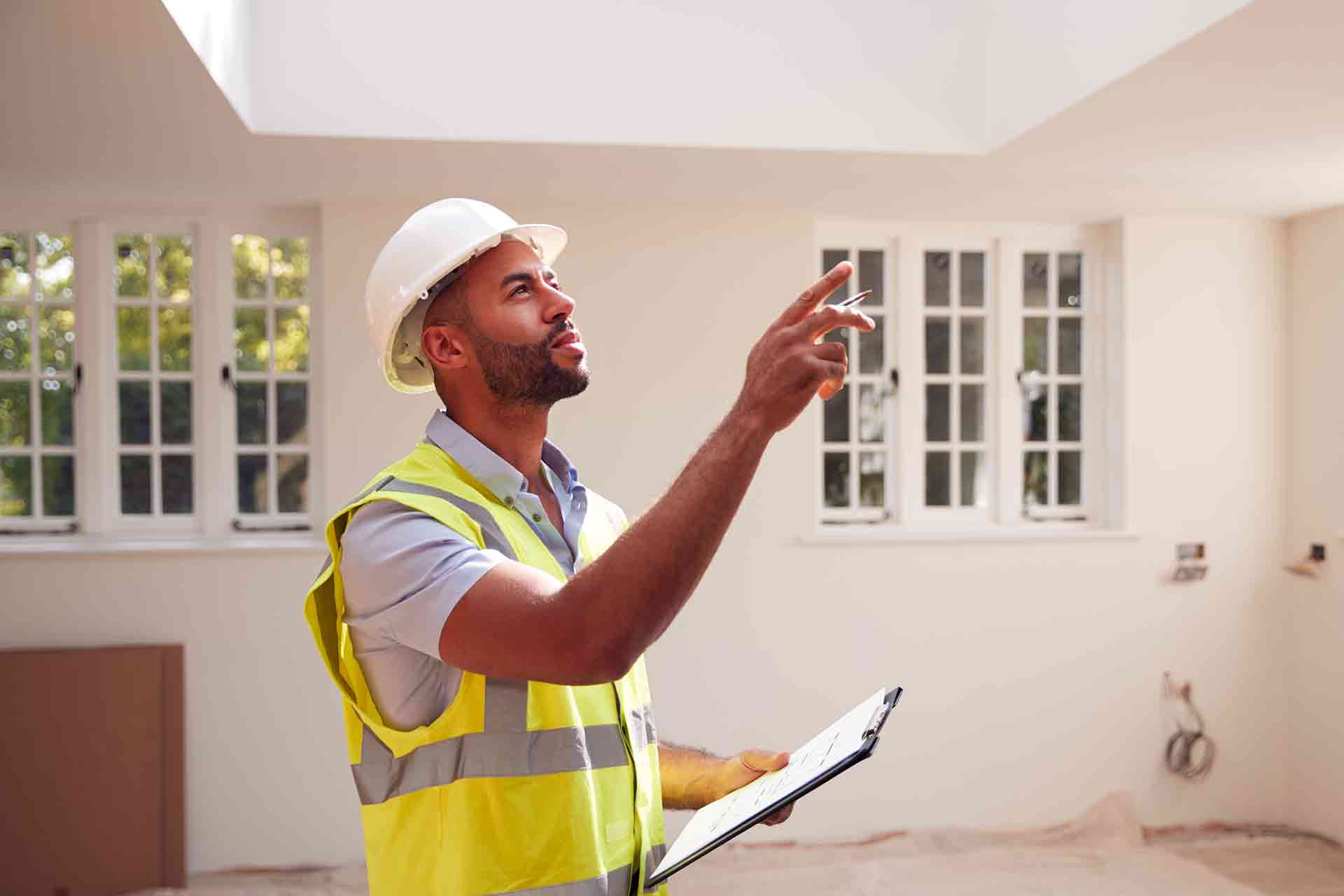 Surveyor in a hard hat and hi-vis vest holding a clipboard and pointing up at a ceiling