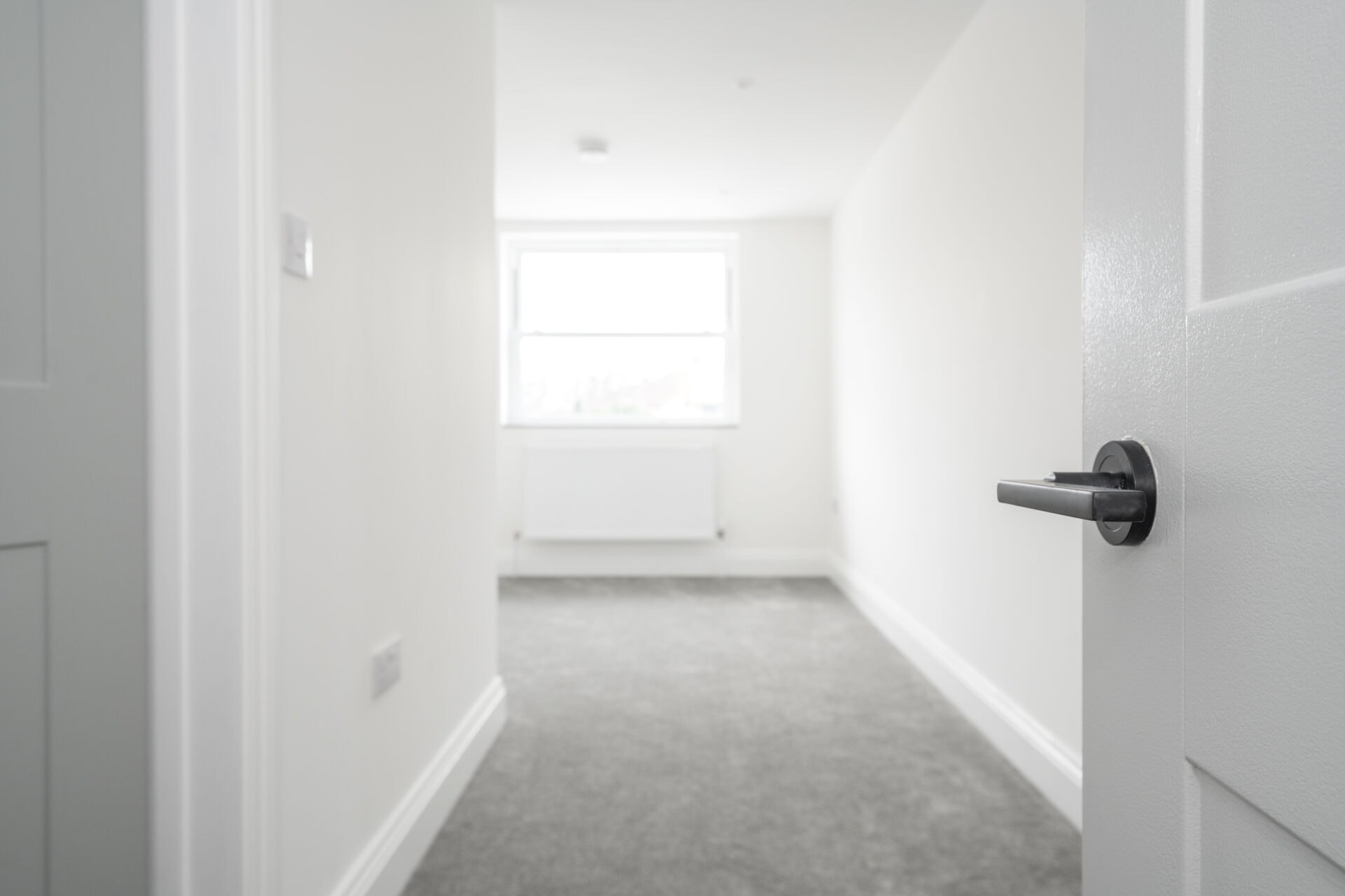 Empty white bedroom with a sash window, radiator and grey carpet, seen past an open door with a black lever handle